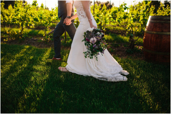 Groom and bride with bouquet walking past grape vines.
