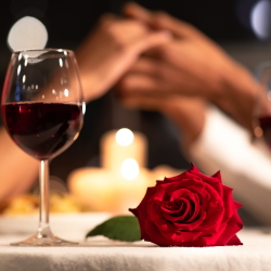 A glass of red wine and a rose with holding hands in the background.