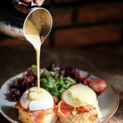 Pouring hollandaise over eggs benedict with a salad in the foreground.